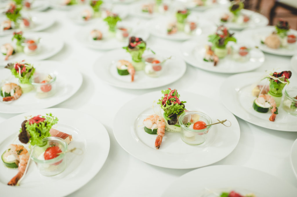 Mehrere weiß gedeckte Teller mit dekorativ angerichteten Vorspeisen, darunter Garnelen, Gurke, Salat und kleine Gläschen mit Tomate und Mozzarella – vorbereitet für eine Veranstaltung oder ein Buffet.